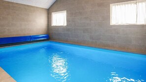 An indoor swimming pool with a blue tiled floor and walls, two windows with white curtains, and a blue safety mat along the edge.