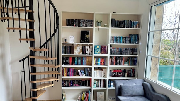 A room with a spiral staircase, a large bookshelf filled with books, a grey sofa, and a large window. - Pleasant family home near Pézenas (Caux)