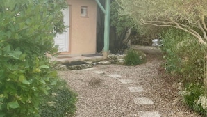A house with a gravel driveway leading to a white door, surrounded by greenery and trees. - Pleasant family home near Pézenas (Caux)
