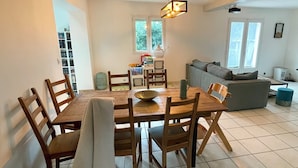 A dining area with a wooden table and chairs, a hanging light fixture, and a view of a living room with a sofa and a window. - Pleasant family home near Pézenas (Caux)