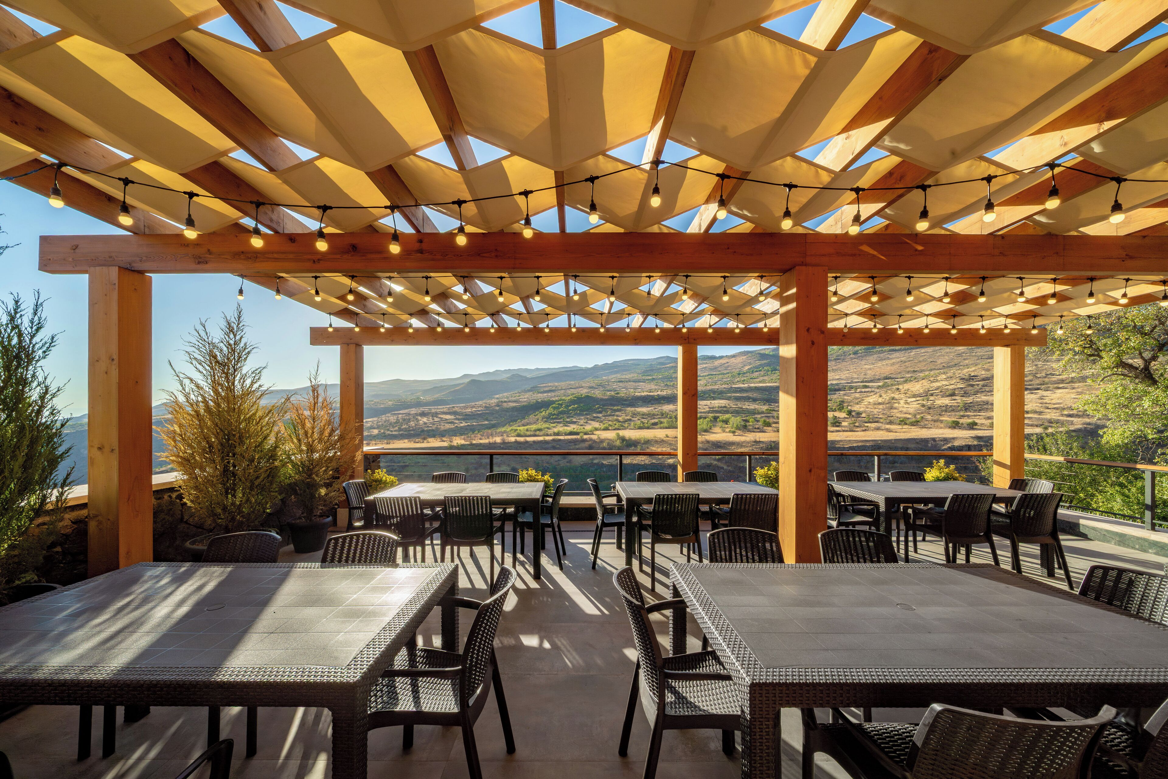 An outdoor patio with tables and chairs, a wooden pergola with string lights, and a scenic view of mountains.