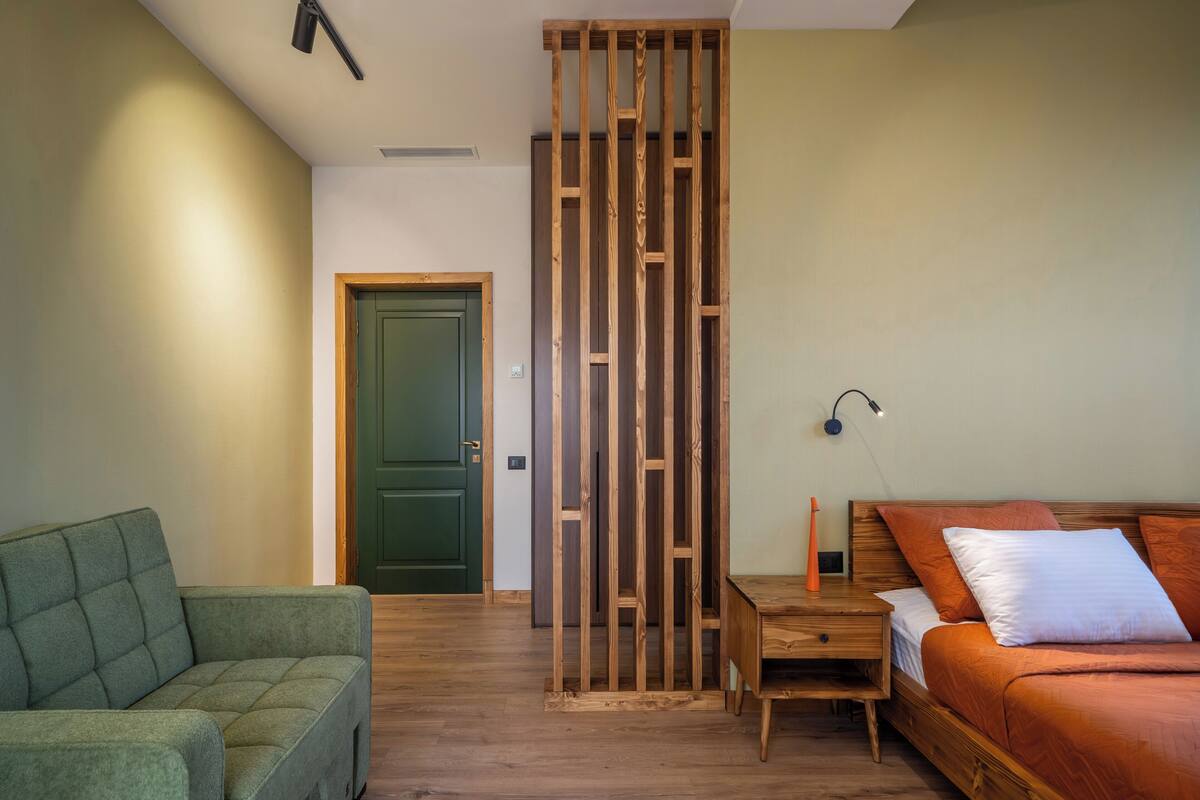 A room with a green sofa, a wooden bed with orange bedding, a wooden partition, and a nightstand with a lamp.