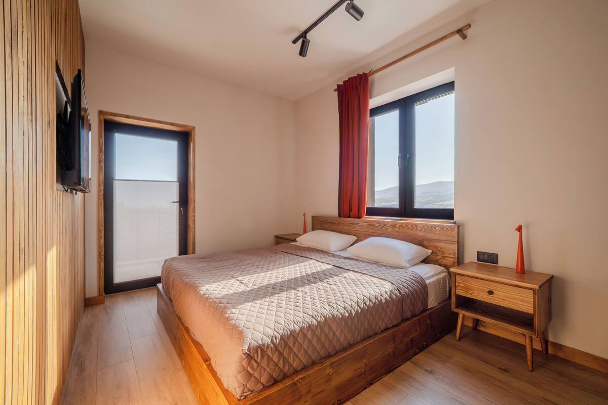 A bedroom with a wooden bed, a nightstand, a TV mounted on the wall, and a window with curtains.