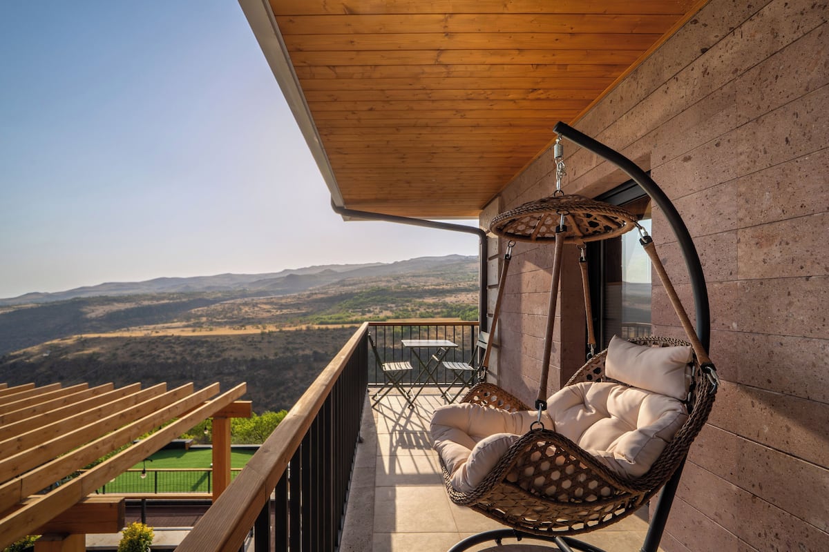A balcony with a hanging chair, a table, and a view of the countryside.