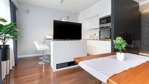 A modern kitchen with a flat-screen TV, white cabinetry, and a dining area with a wooden table and a potted plant. - Modern Apartment | Private Garden and Parking (Gdańsk)