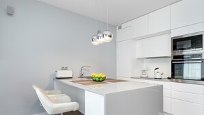 A modern kitchen with white cabinetry, a central island with a bowl of fruit, and a bar stool. - Modern Apartment | Private Garden and Parking (Gdańsk)
