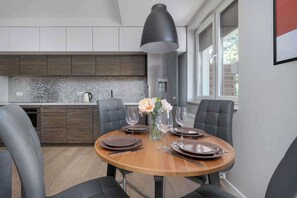 A modern dining area with a round wooden table set for four, surrounded by black upholstered chairs. A large pendant light hangs above the table, and a vase with flowers is placed in the center. The background features a kitchen with dark wooden cabinets and a tiled backsplash.