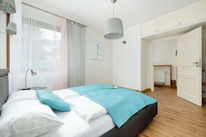 A bedroom with a bed, a bedside lamp, a window with sheer curtains, a painting on the wall, and a door leading to another room.