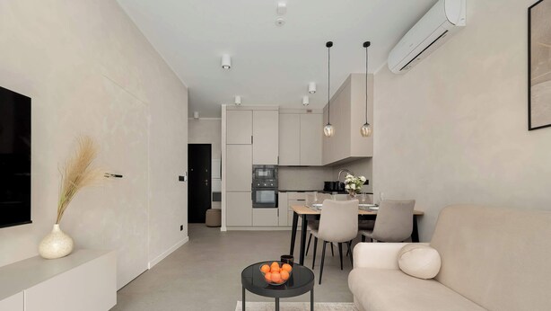 Un salon moderne avec un canapé, une table basse et un téléviseur fixé au mur. On aperçoit une cuisine à l’arrière-plan, avec des placards et un coin repas.