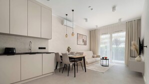 Une cuisine moderne avec des meubles blancs, un coin repas comprenant une table et des chaises, et un salon avec un canapé et de grandes fenêtres.