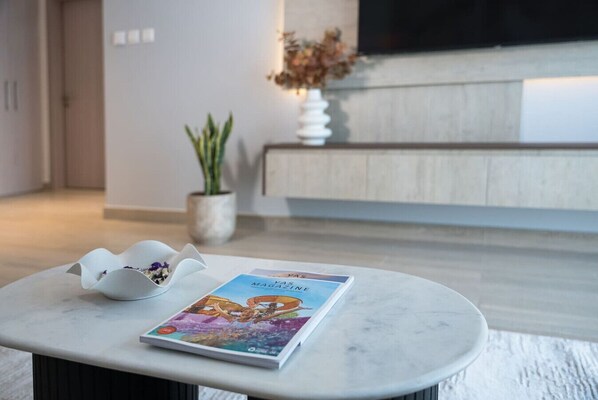 A modern living room with a marble coffee table, a magazine, a white decorative bowl, a potted plant, and a flat-screen TV mounted on the wall.