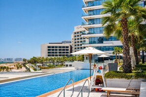 A hotel pool area with a clear view of the sea, palm trees, and a modern building with balconies.