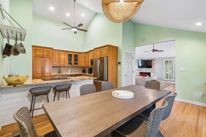 A modern kitchen with wooden cabinets, a central island, and a dining area with a long table and wicker chairs. - IOP Beach House with a Tiki Bar (Isle of Palms)