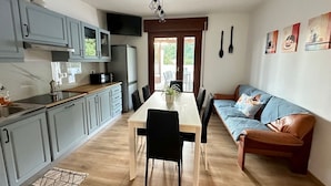 Eine kleine Küche mit grauen Schränken, einer Mikrowelle und einem Kühlschrank. Es gibt einen Esstisch mit Stühlen und eine Couch mit Kissen.
