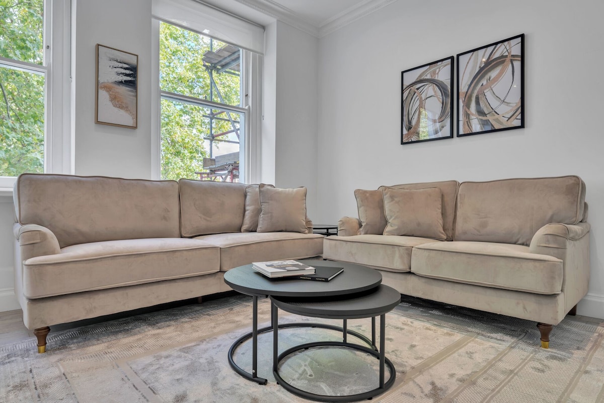 A corner living room with two beige sofas, a round coffee table, and abstract wall art.