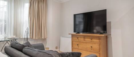 A modern living room with a flat-screen TV, a sofa, a wooden cabinet, and a dining area with a glass table and chairs.