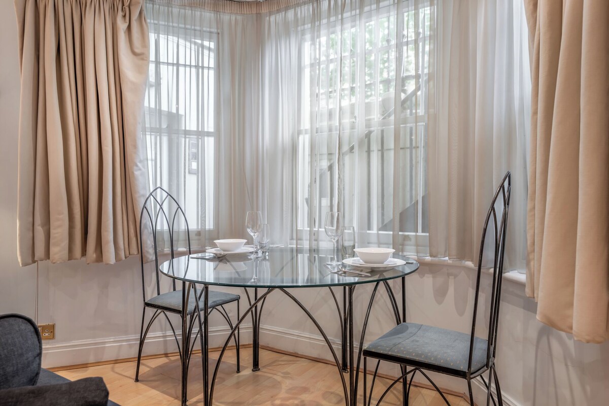 A glass-top dining table with four chairs, a window with sheer curtains, and a staircase visible through the window.
