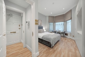 A bedroom with a bed, bedside table, chair, and a window with curtains. - The Beaufort Apartments (London)