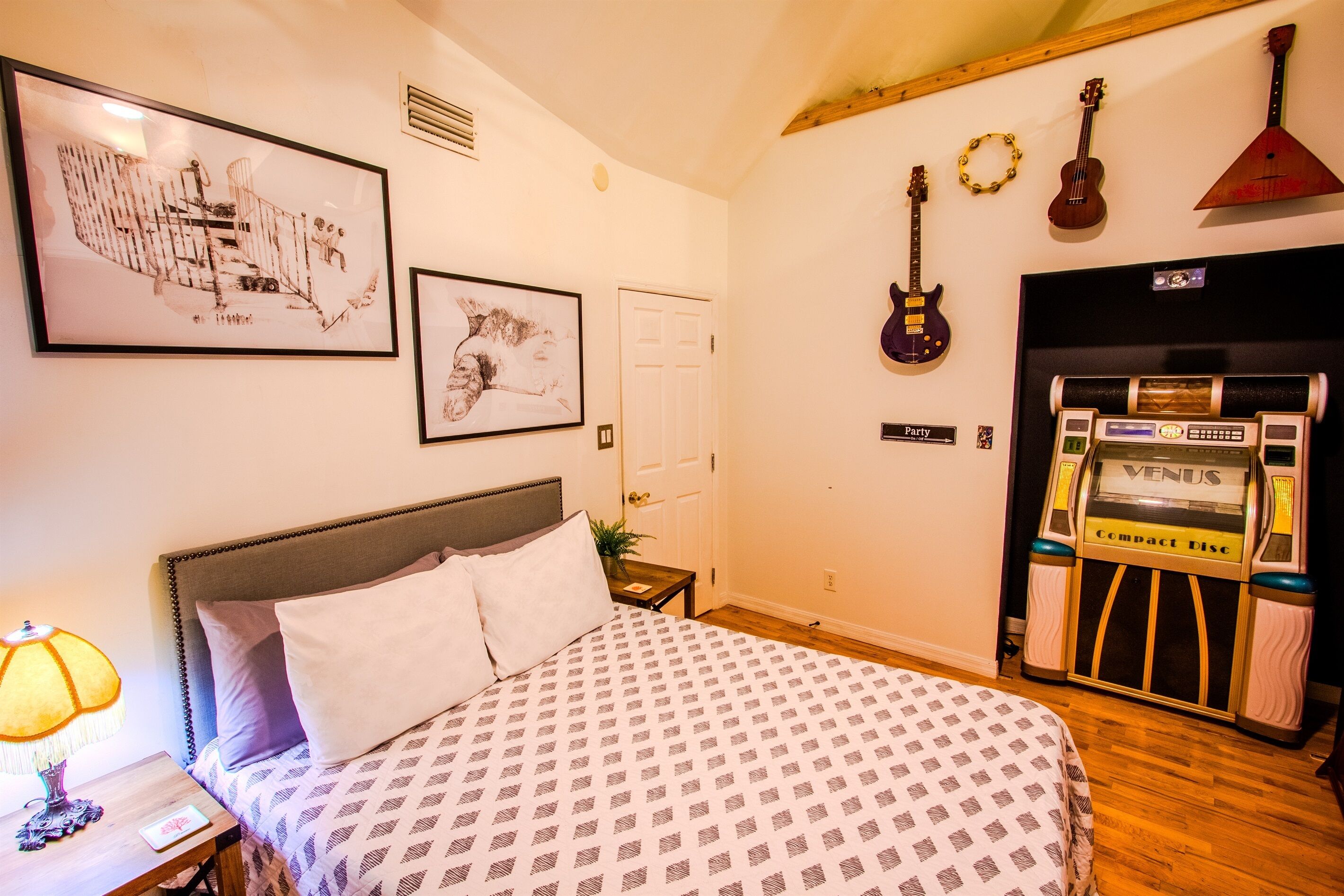 A bedroom with a bed, a nightstand, a lamp, framed artwork, a door, and a jukebox.
