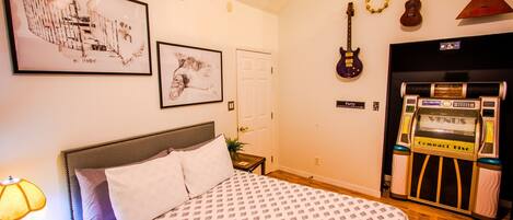 A bedroom with a bed, a nightstand, a lamp, framed artwork, a door, and a jukebox.