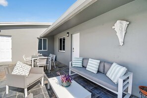 A modern outdoor seating area with a sofa, chairs, and a table, featuring a stone floor and a white wall with a decorative element.