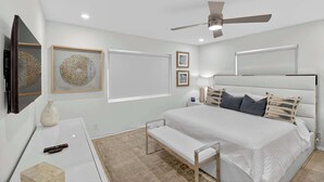 Un dormitorio moderno con una cama grande, una mesita de noche, un banco, un ventilador de techo y obras de arte colgadas en la pared.