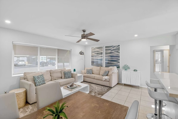 A modern living room with two sofas, a coffee table, a dining area with a table and chairs, and a large window with a view of the street.