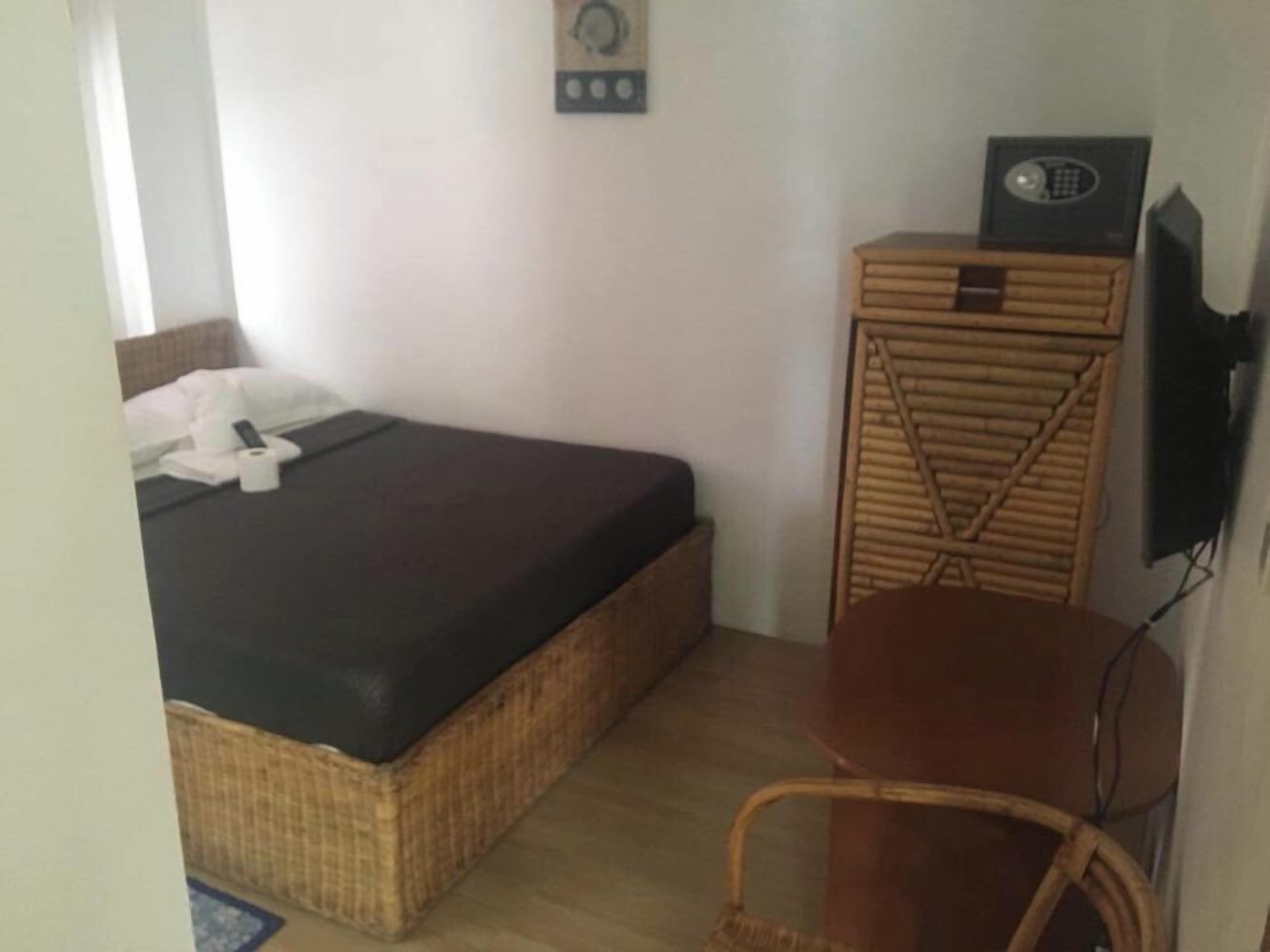 A small room with a bed, a wicker chair, a television, and a wall-mounted control panel.