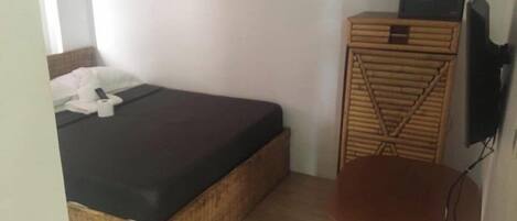 A small room with a bed, a wicker chair, a television, and a wall-mounted control panel.