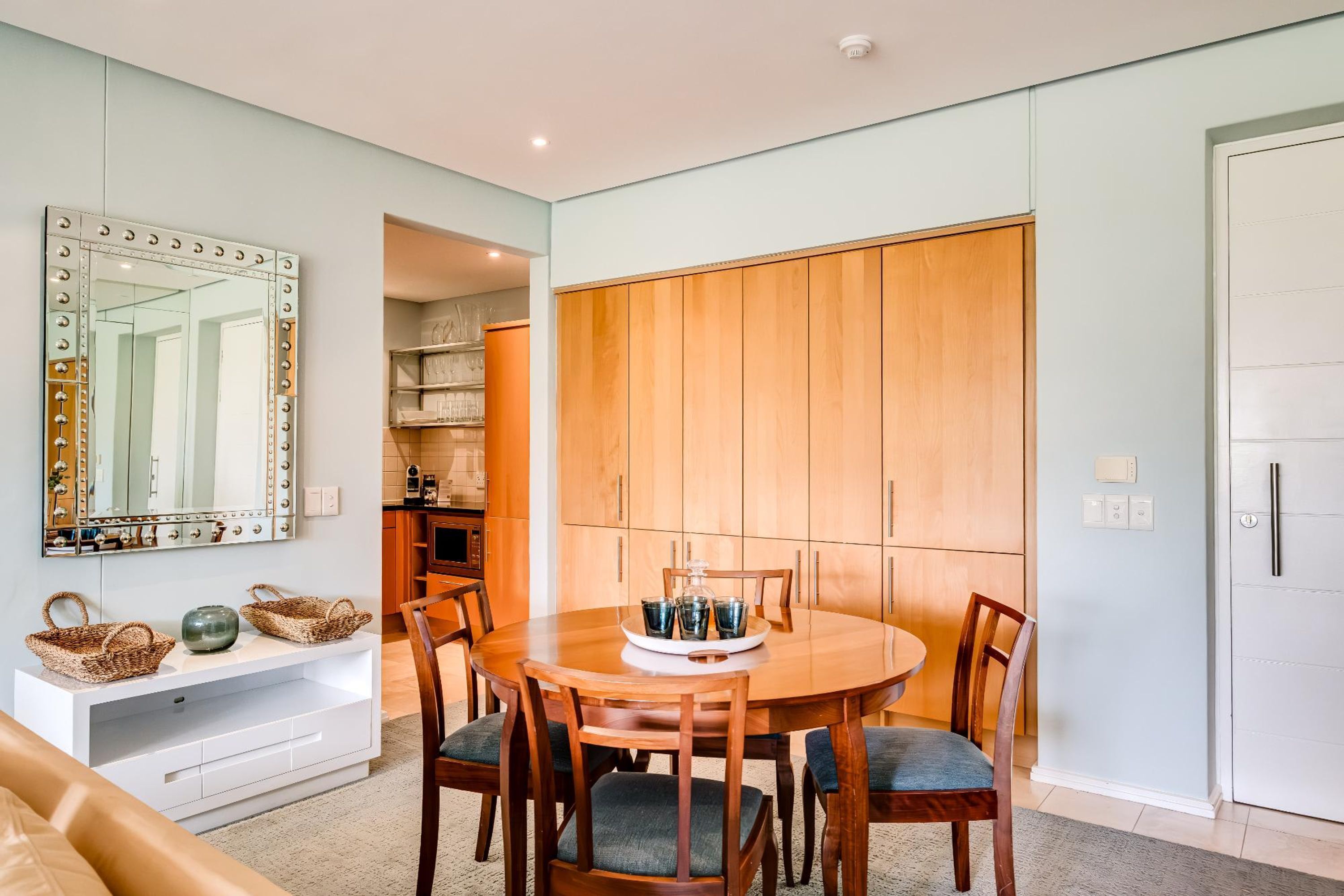 Un comedor con una mesa redonda de madera, sillas, una consola blanca y un espejo grande con luces.