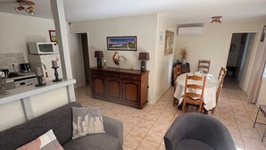 Un salon comprenant un canapé, un fauteuil et une table basse. On y trouve également un coin cuisine avec un four à micro-ondes et une cuisinière. Une table à manger avec des chaises est visible au fond.