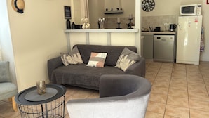 Un salon avec un canapé gris, une petite table ronde et un fauteuil gris. On aperçoit une cuisine à l’arrière-plan, avec un four à micro-ondes, un réfrigérateur et un évier.