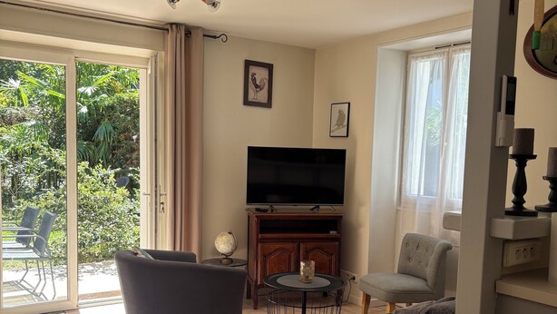 Un salon comprenant un canapé, un téléviseur et une petite table. Une porte-fenêtre coulissante donne accès à un espace extérieur aménagé avec des chaises et de la verdure.