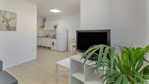 Ett modernt vardagsrum med en platt-TV, ett vitt soffbord, en grå soffa, ett kök i bakgrunden och en tavla på väggen.