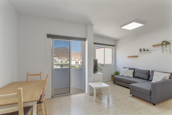 A modern living room with a sliding glass door leading to a balcony, a grey sofa, a white table, and a wooden dining table.