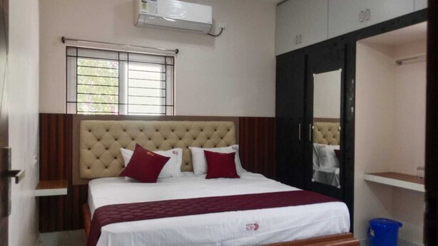 A bedroom with a tufted headboard, a bed with white bedding and red pillows, a window with blinds, and a wardrobe with a mirror.
