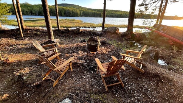 Een kampvuur met een pan, omringd door houten stoelen, in een bebost gebied met een meer op de achtergrond.