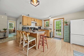 A modern kitchen with wooden cabinets, a central island with stools, a refrigerator, and a view through the door. - Rustic and Stylish Cabin on Stunning Serene Pond (Blue Hill)