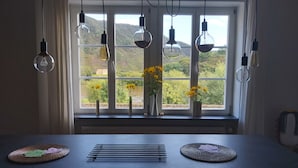 A dining table with a view of a river and hills through large windows, with hanging light bulbs and vases with yellow flowers.
