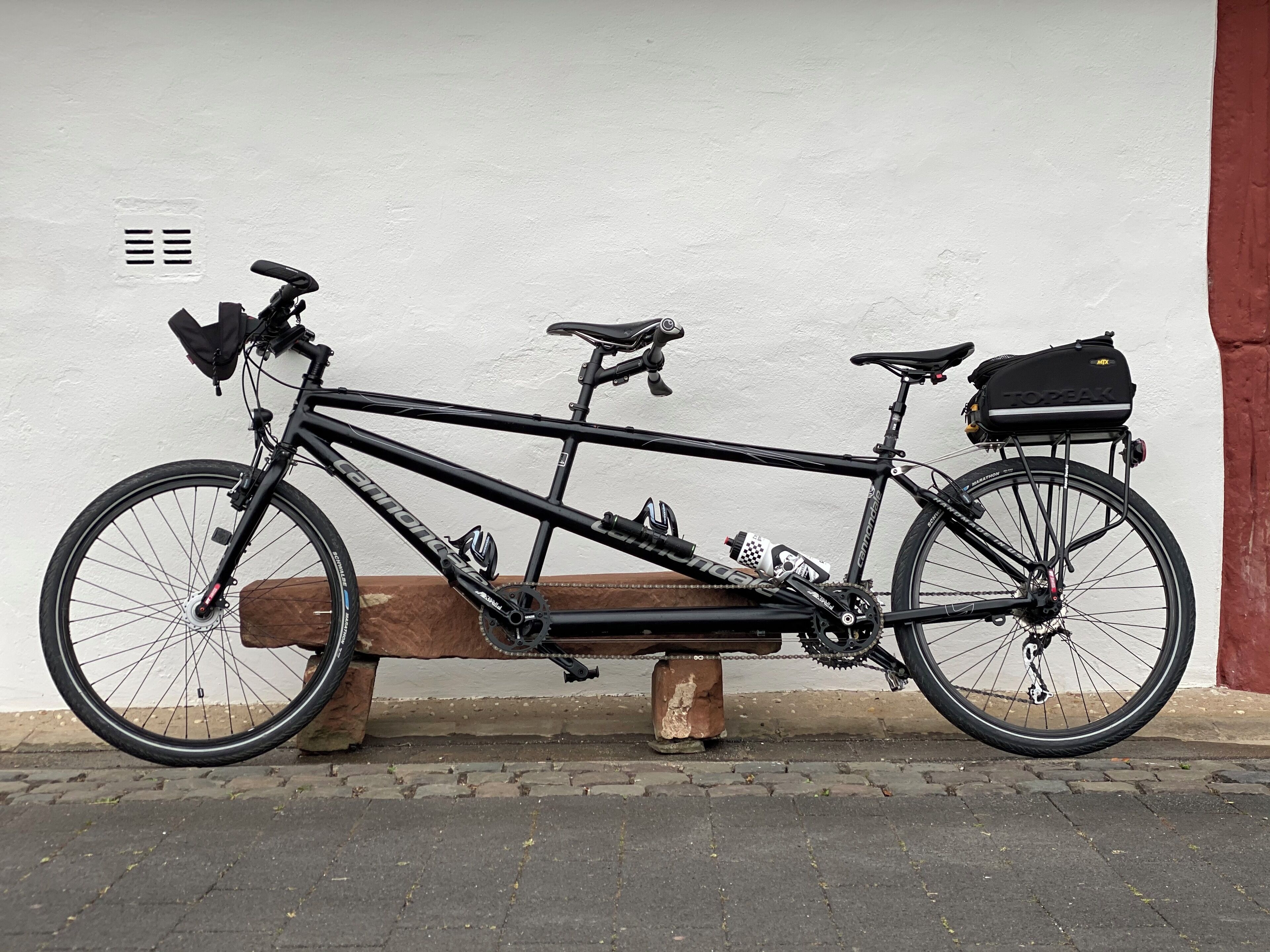 A black bicycle with a saddlebag and water bottle, parked on a bench against a white wall.