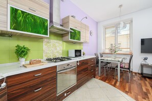 A modern kitchen with wooden cabinets, a built-in oven, and a microwave. There is a dining table with chairs and a small side table with a lamp.