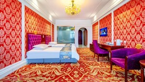 Habitación de hotel con una cama grande, un escritorio y una silla. Se aprecia una araña colgante sobre la cama y un ventanal grande al fondo.
