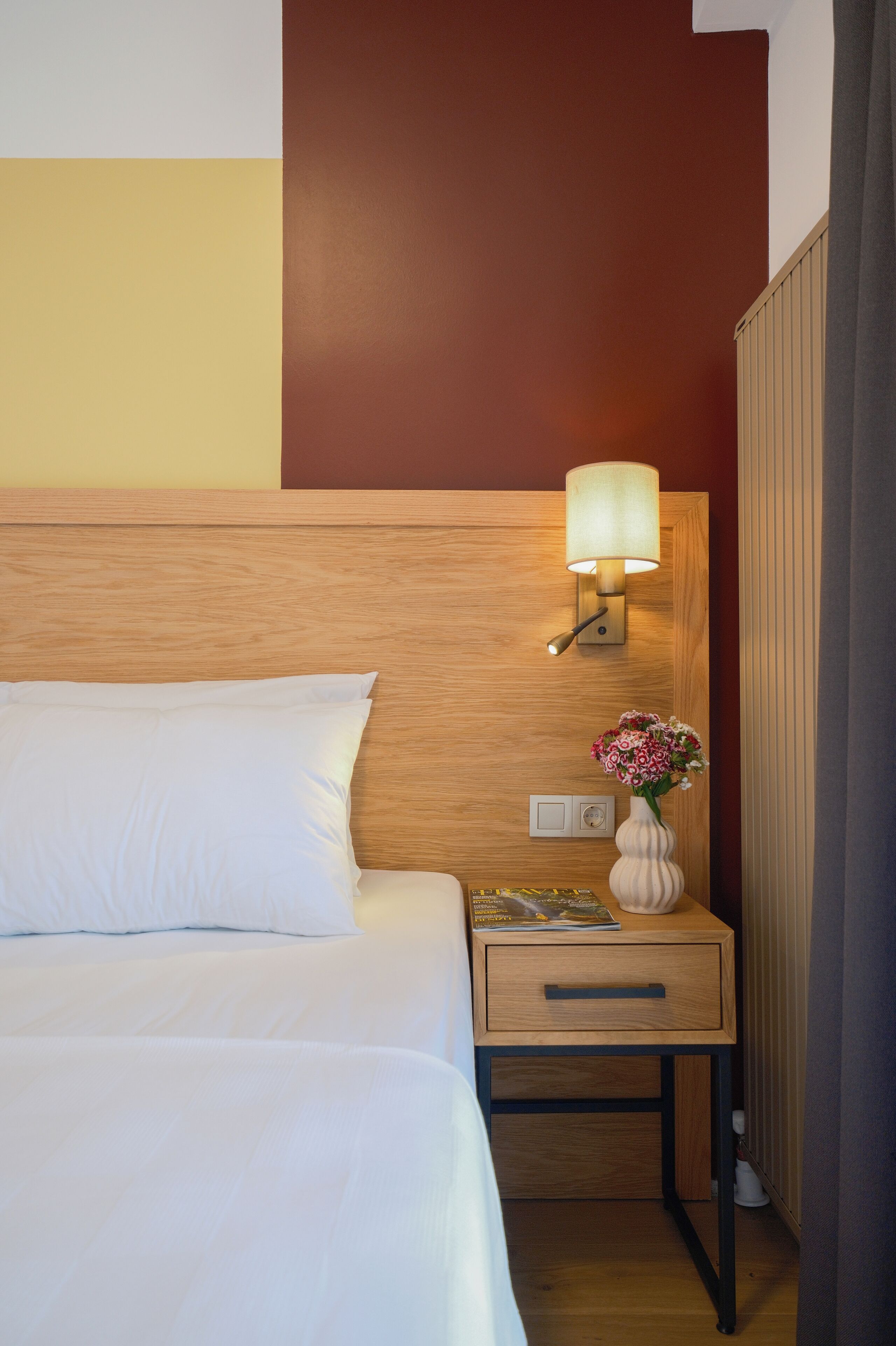 A hotel room with a wooden headboard, a bedside lamp, a nightstand with a vase of flowers, and a drawer.