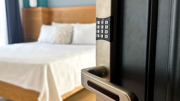 A modern hotel room with a bed, a door with a numeric keypad lock, and a wooden headboard.