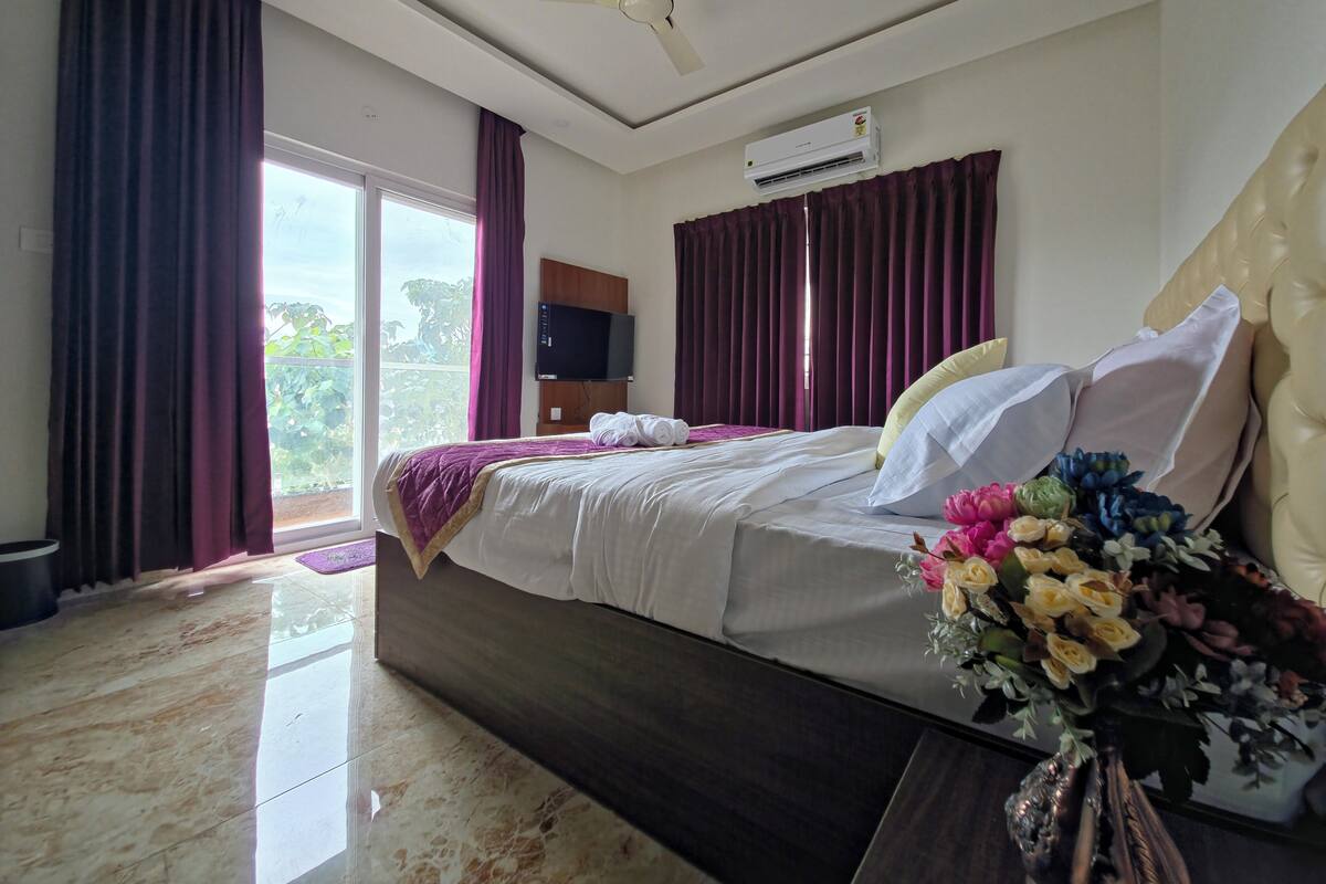 A hotel room with a bed, a ceiling fan, an air conditioning unit, a TV, and a large window with curtains.