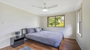 Un dormitorio con cama, mesita de noche, lámpara, ventilador de techo y ventana con vistas a zonas verdes.
