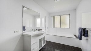 Une salle de bain moderne avec un meuble-lavabo blanc, un grand miroir, une baignoire et une fenêtre.