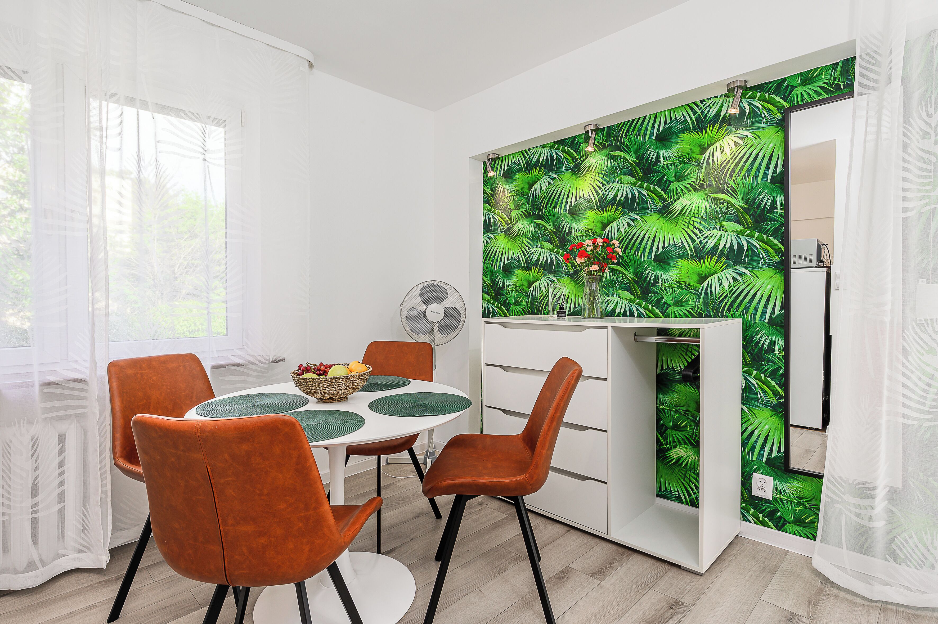 A modern dining area with a white table, orange chairs, and a green leaf-patterned wall.
