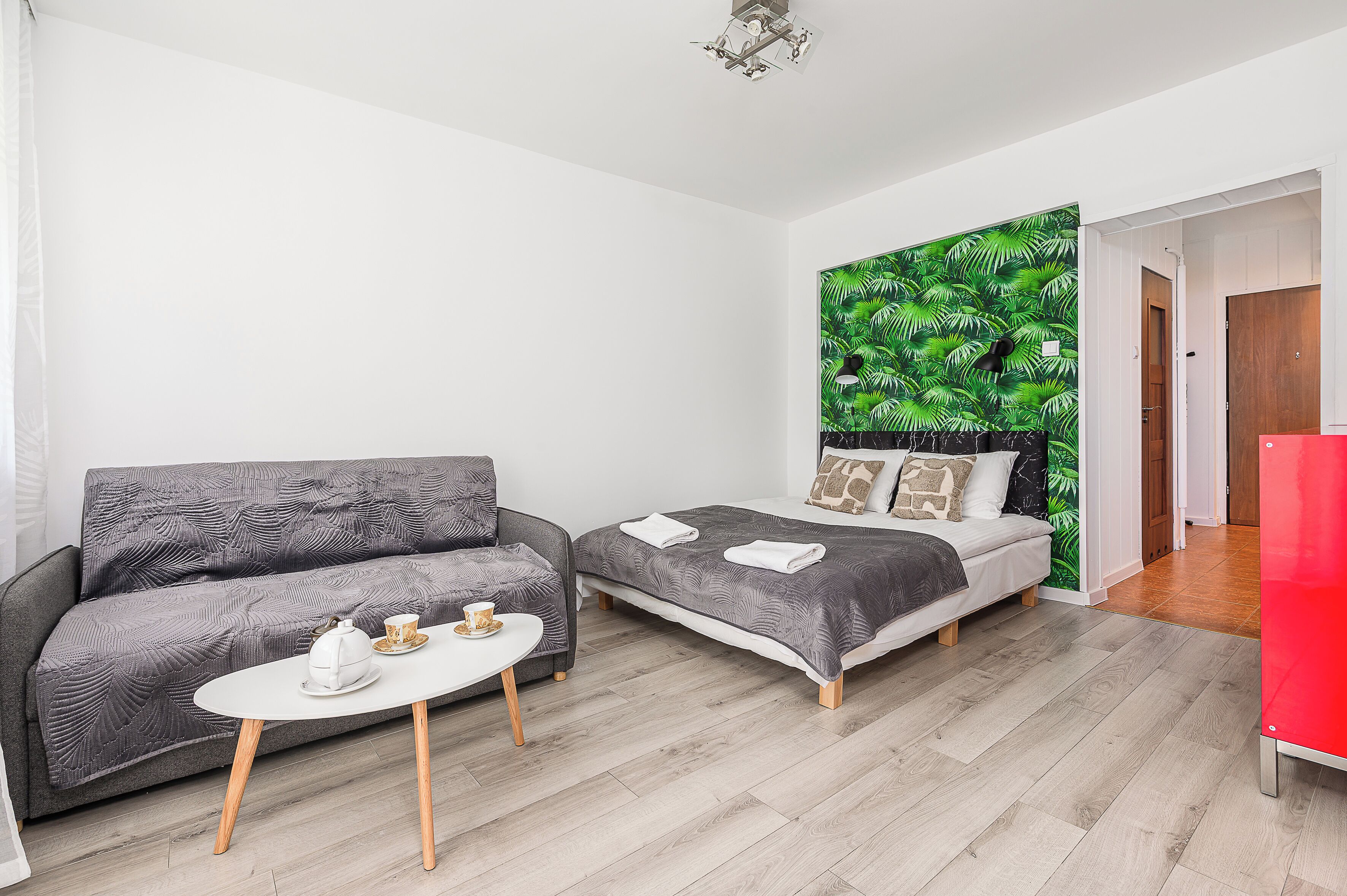A modern room with a grey sofa, a bed with a grey bedspread, a white coffee table with a teapot and cup, and a green leaf-patterned wall.