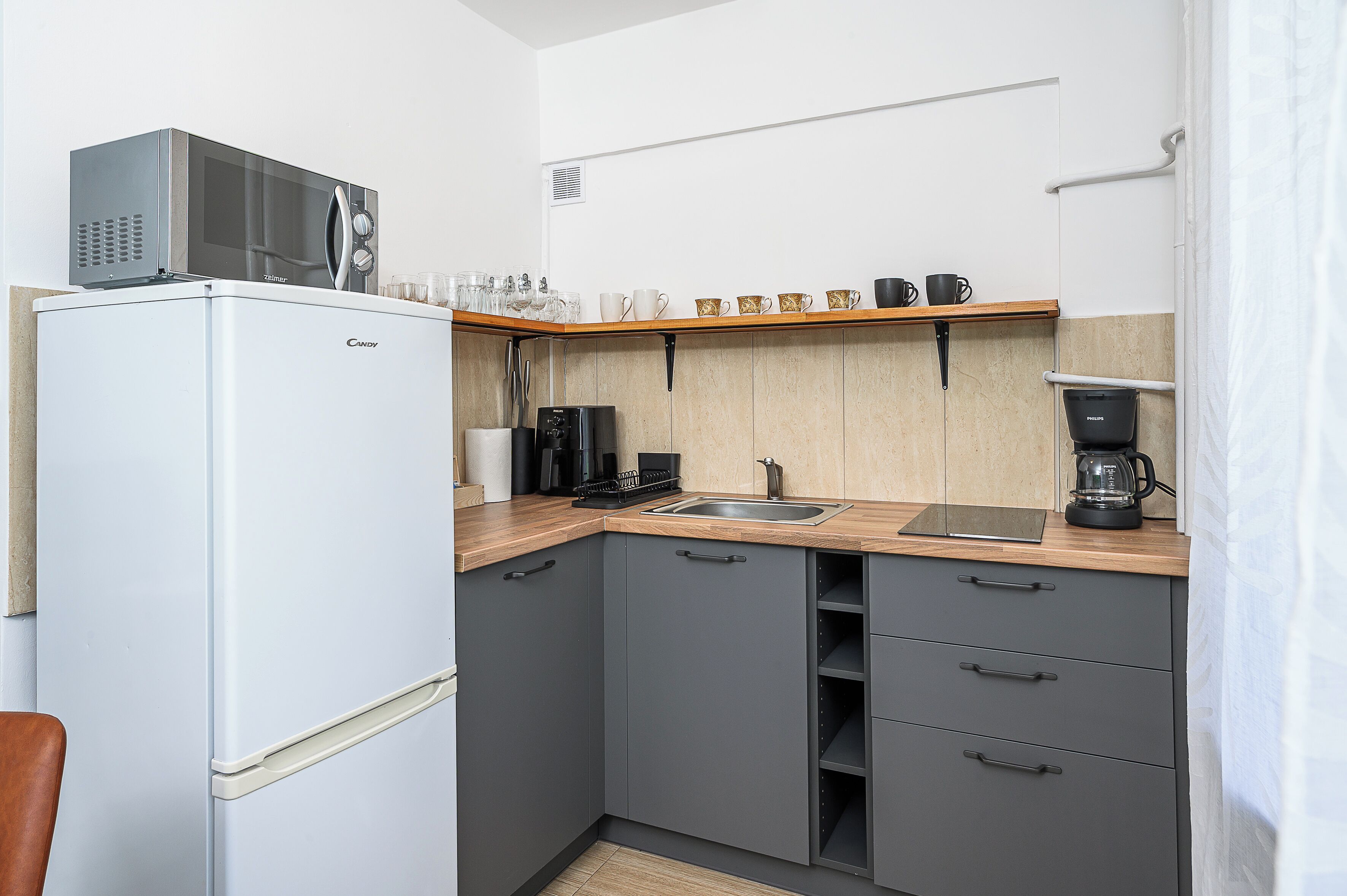 A compact kitchen with a refrigerator, microwave, coffee maker, and a shelf with cups and glasses.